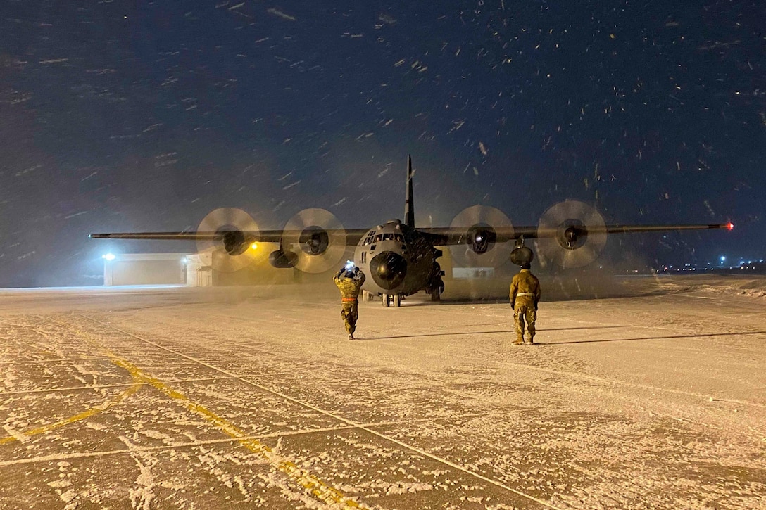Two service members guide an aircraft as snow falls at night.