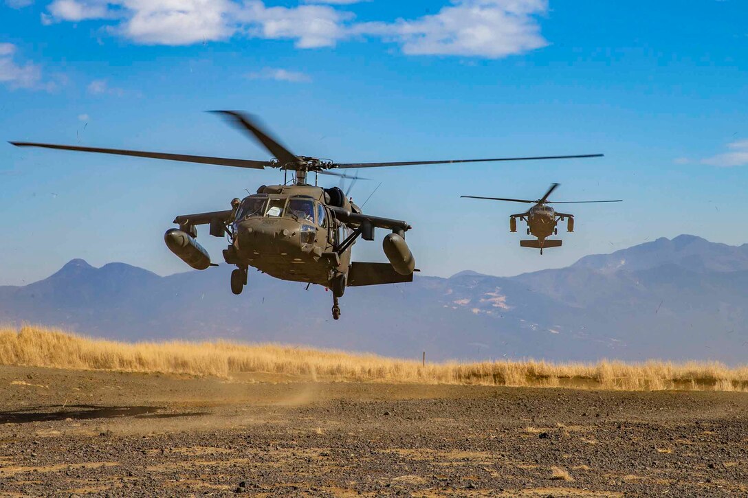 Two helicopters fly over desert terrain; a mountain range can be seen behind.