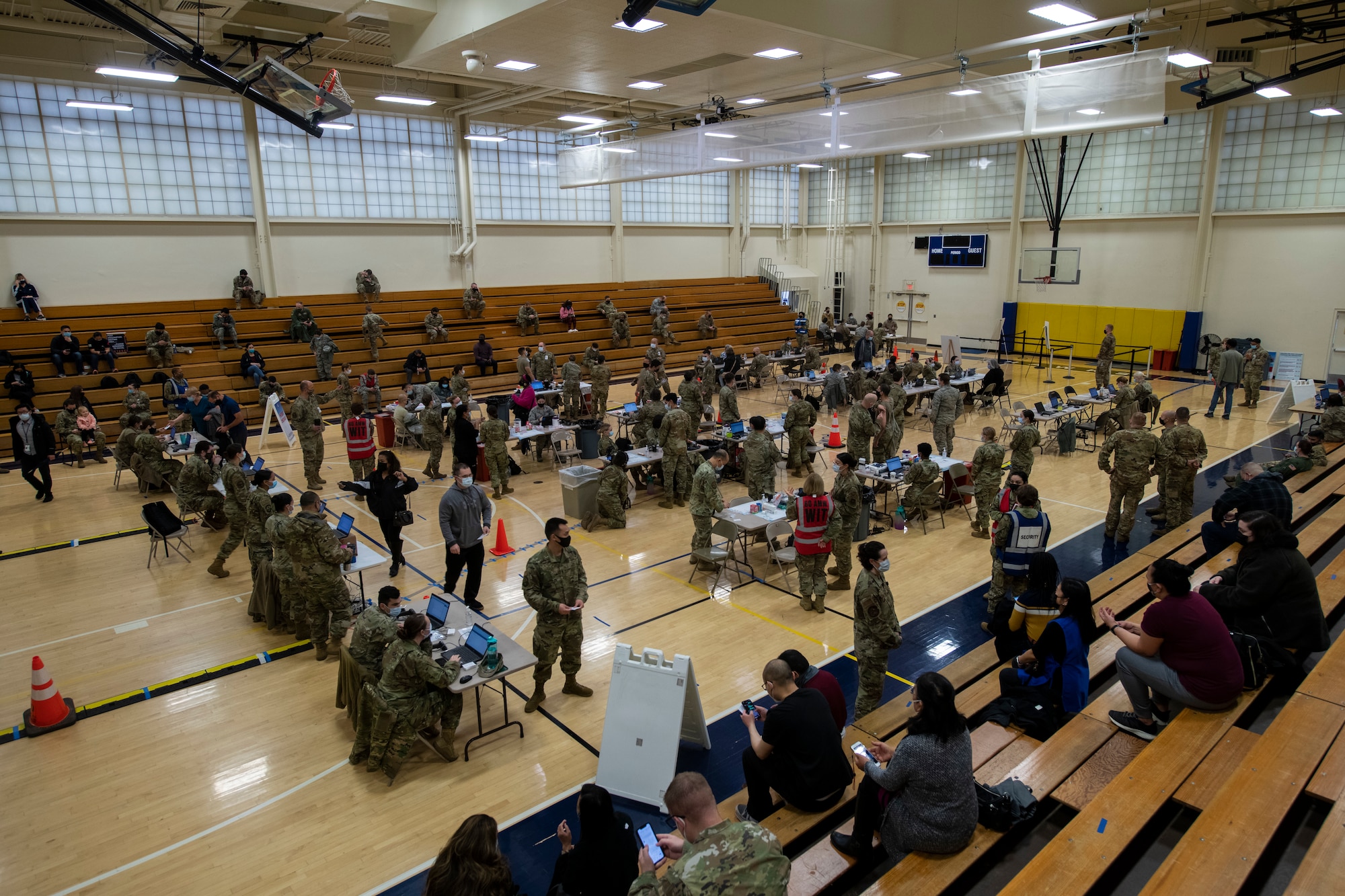 A large gymnasium has many people administering vaccines to people while the bleachers are sporadically covered with various people.