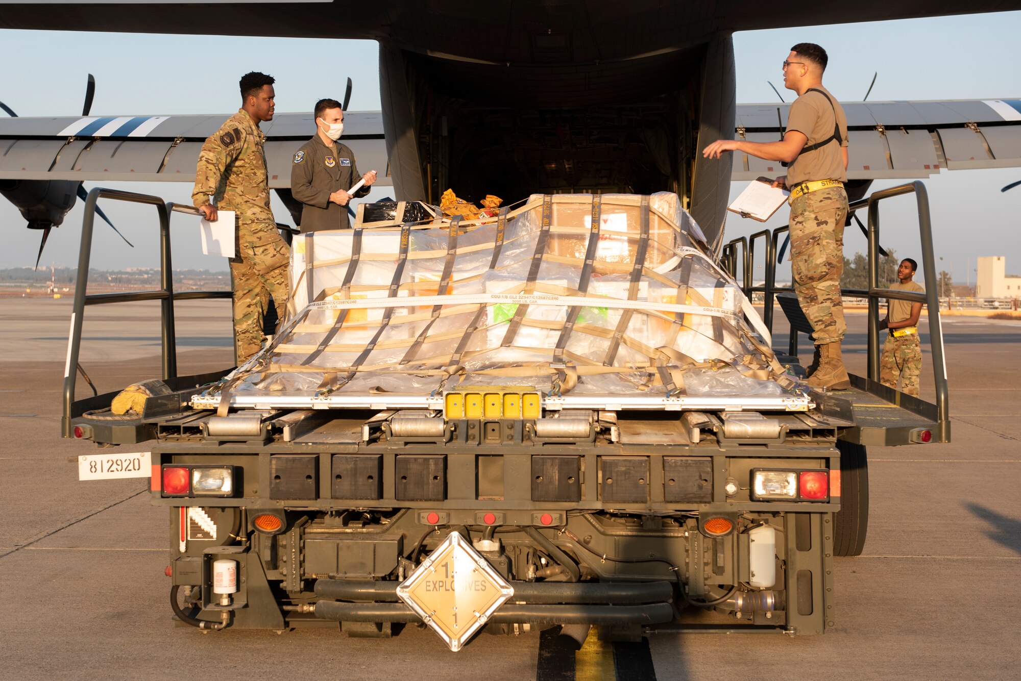 Three Airmen stand on a cargo loading truck, which is unloading from the back of a large propeller cargo aircraft.