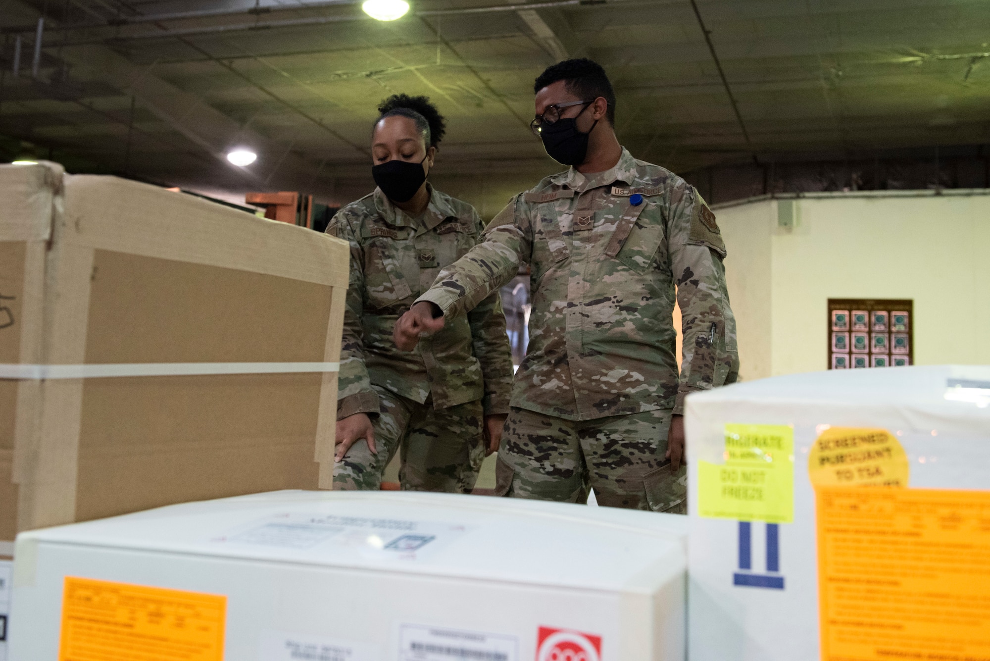 Two Airmen stand in front of small cargo boxes and inspect the labels.