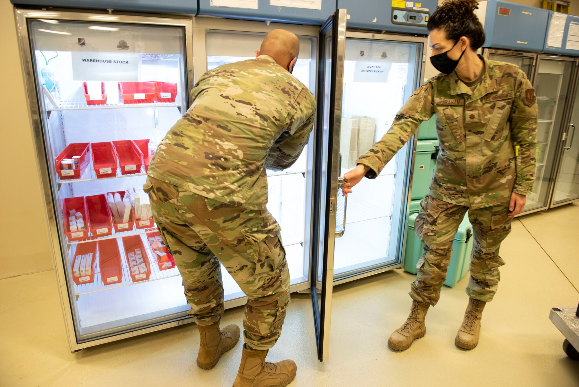 An female Major holds open a refrigerator door for a male Airman to put vials of medicine inside.