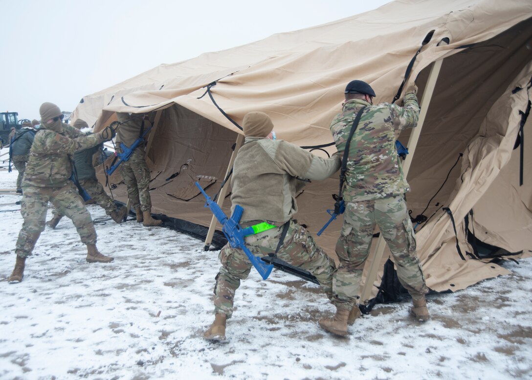 A group of Airmen in a snowy area pulls several ropes at the same time, in order to lift the middle of the tent.