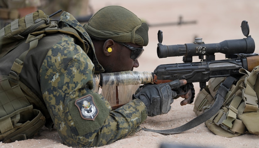 A soldier lays on the ground and aims a rifle.
