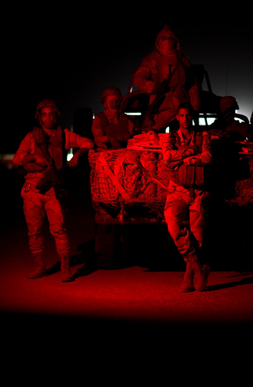 Military personnel stand at the rear of a military vehicle at night.