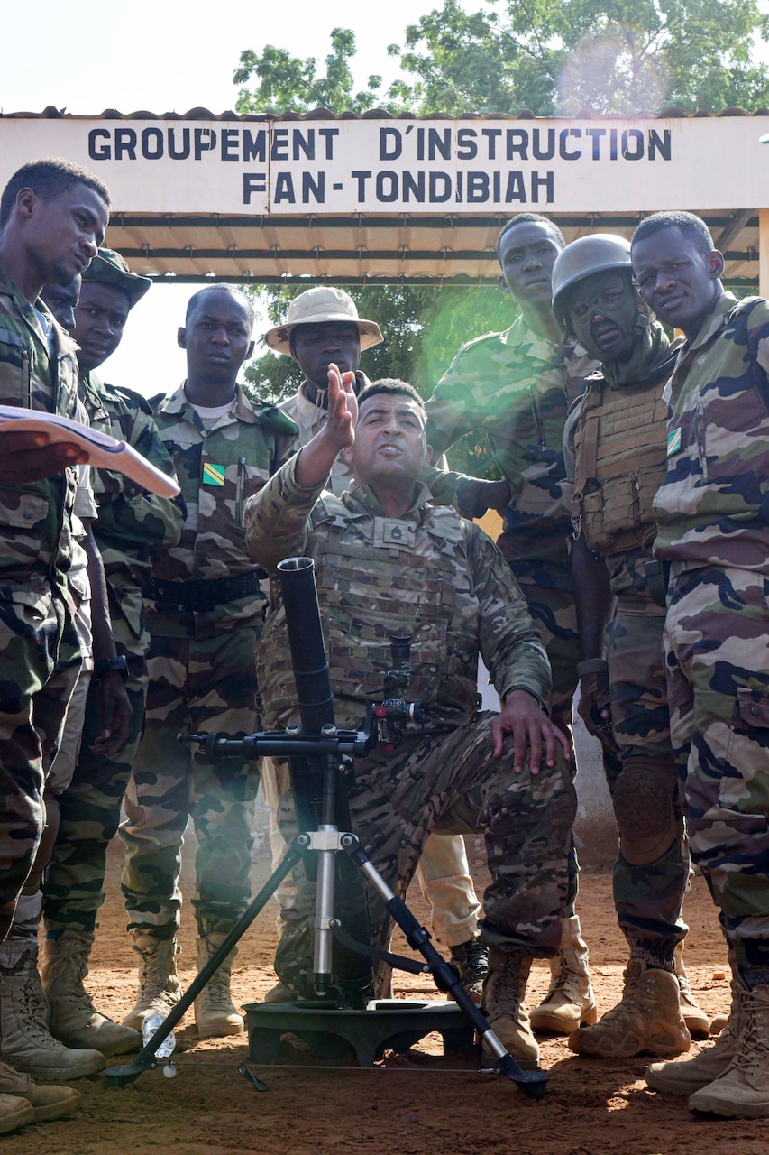 A soldier kneels in front of a weapon while soldiers stand around him.