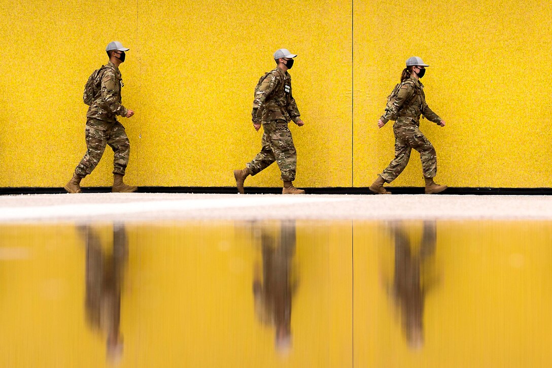 Three Air Force cadets walk in a line next to a yellow wall.