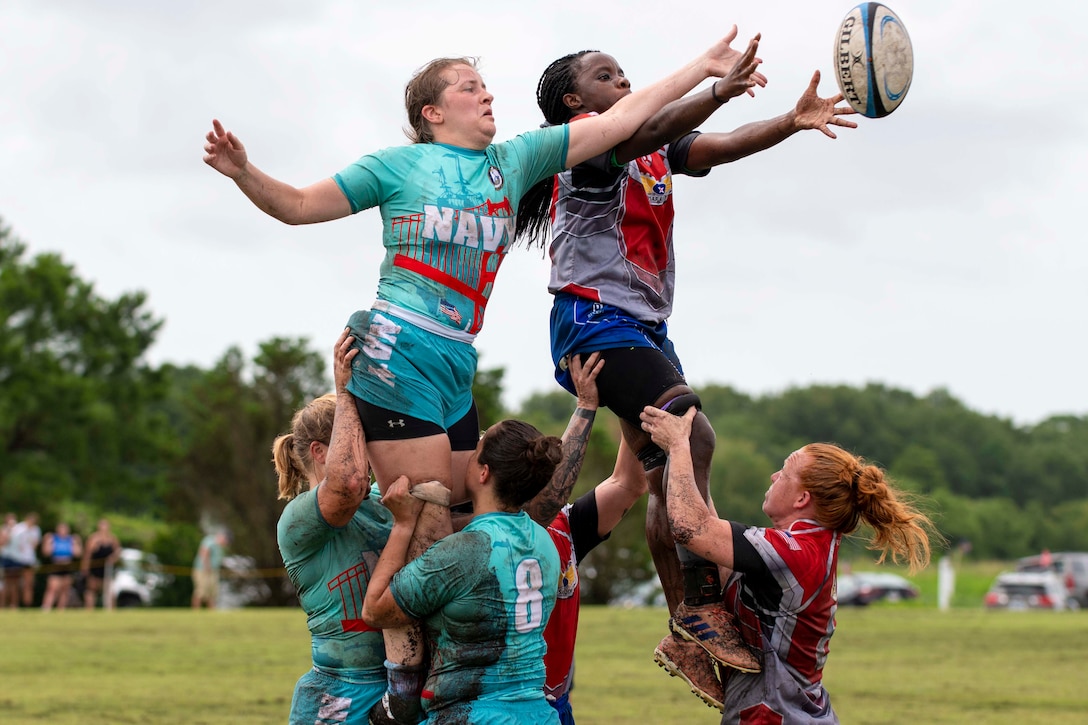 Two airmen and two sailors lift two others up as they try to grab a rugby ball.