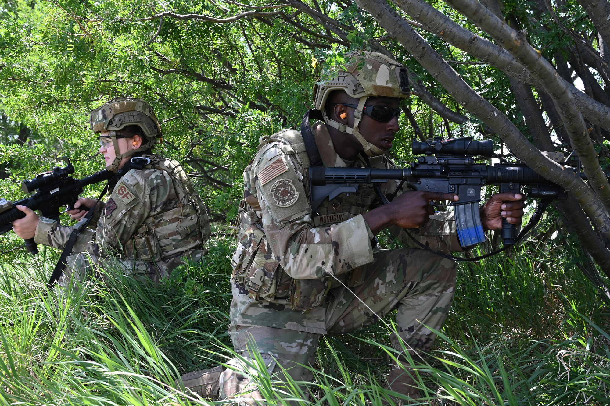 Two North Dakota Air National Guard members of the 219th Security Forces Squadron in uniform crouch near some bushes with training M4 rifles during a training exercise at the Minot Air Force Base, N.D., June 24, 2021.