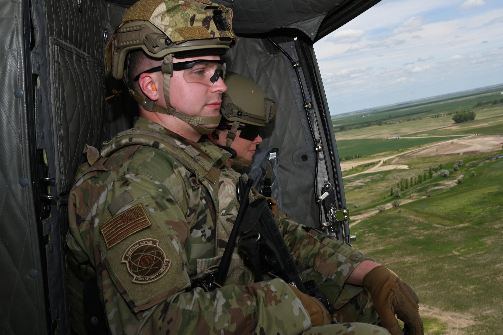 Two North Dakota Air National Guard 219 Security Forces Squadron members wearing military uniforms sit in a UH-1N Huey helicopter, looking out an open door, as they fly over a rural area near the Minot Air Force Base, N.D., June 24, 2021.