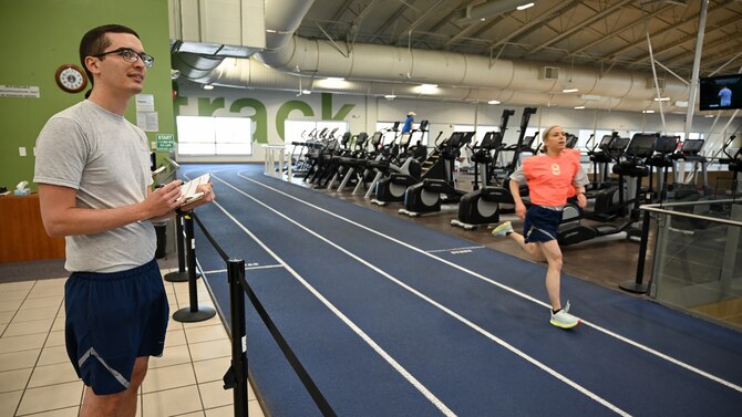 Staff Sgt. Nicole Ligeza runs on an indoor track while Tech. Sgt. Alexander House records her run time in the foreground.