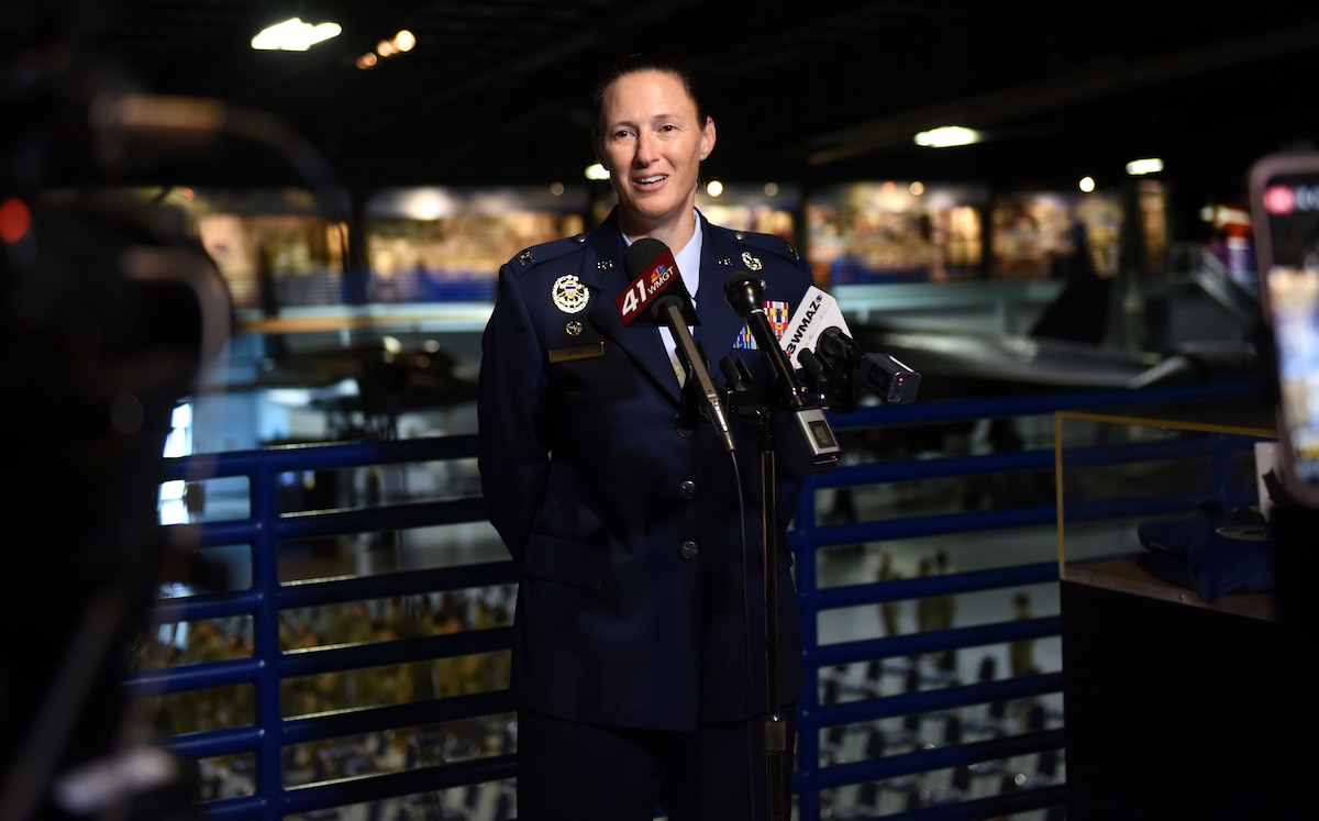 Photo shows Col. Droz standing in front of a mic stand speaking into multiple microphones.