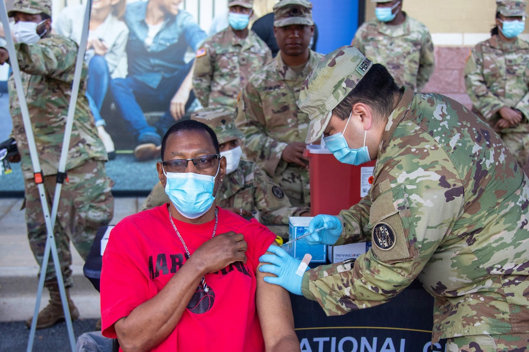 A soldier vaccinates a man as other soldiers stand behind them.