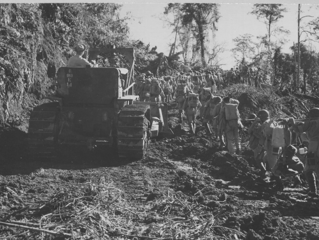 A line of soldiers walks past a large tractor that’s working to clear a dirt road.