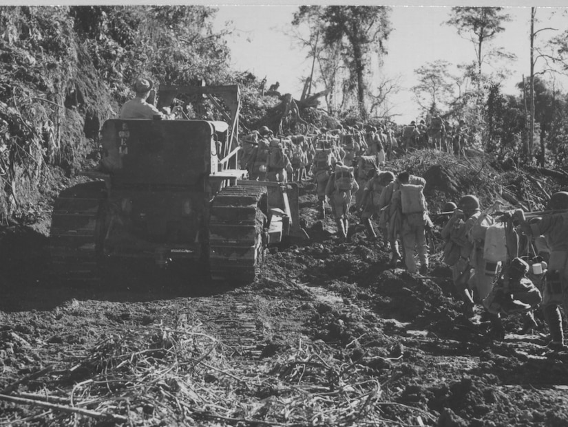 A line of soldiers walks past a large tractor that’s working to clear a dirt road.