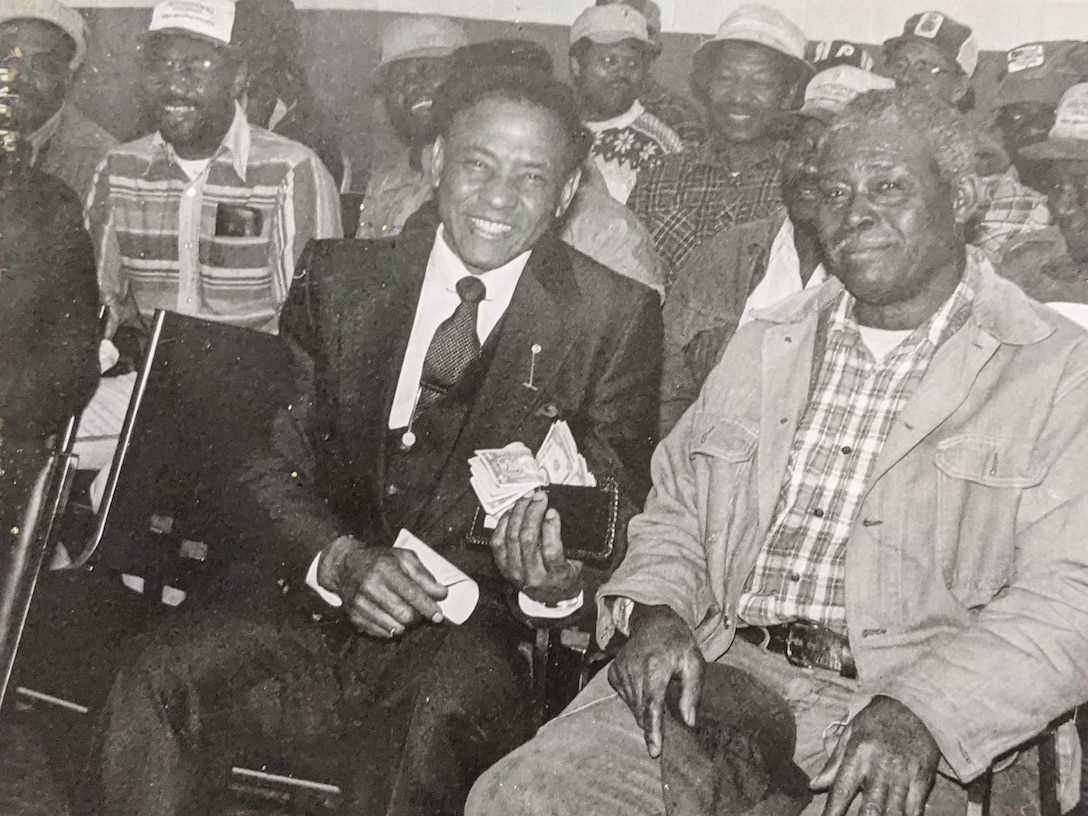 Several men sit grouped together. One smiles while holding up a wallet spilling over with cash.