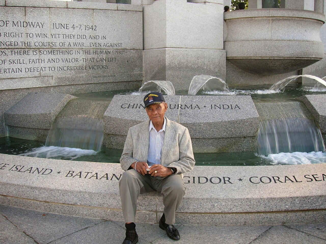 A man wearing a suit and baseball cap sits in front of a water fountain monument.
