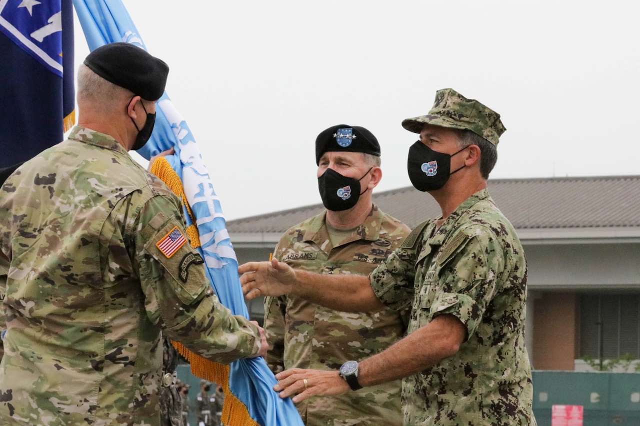 A man in a military uniform holds a flag in his hands and presents it to another man in uniform as a third looks on.