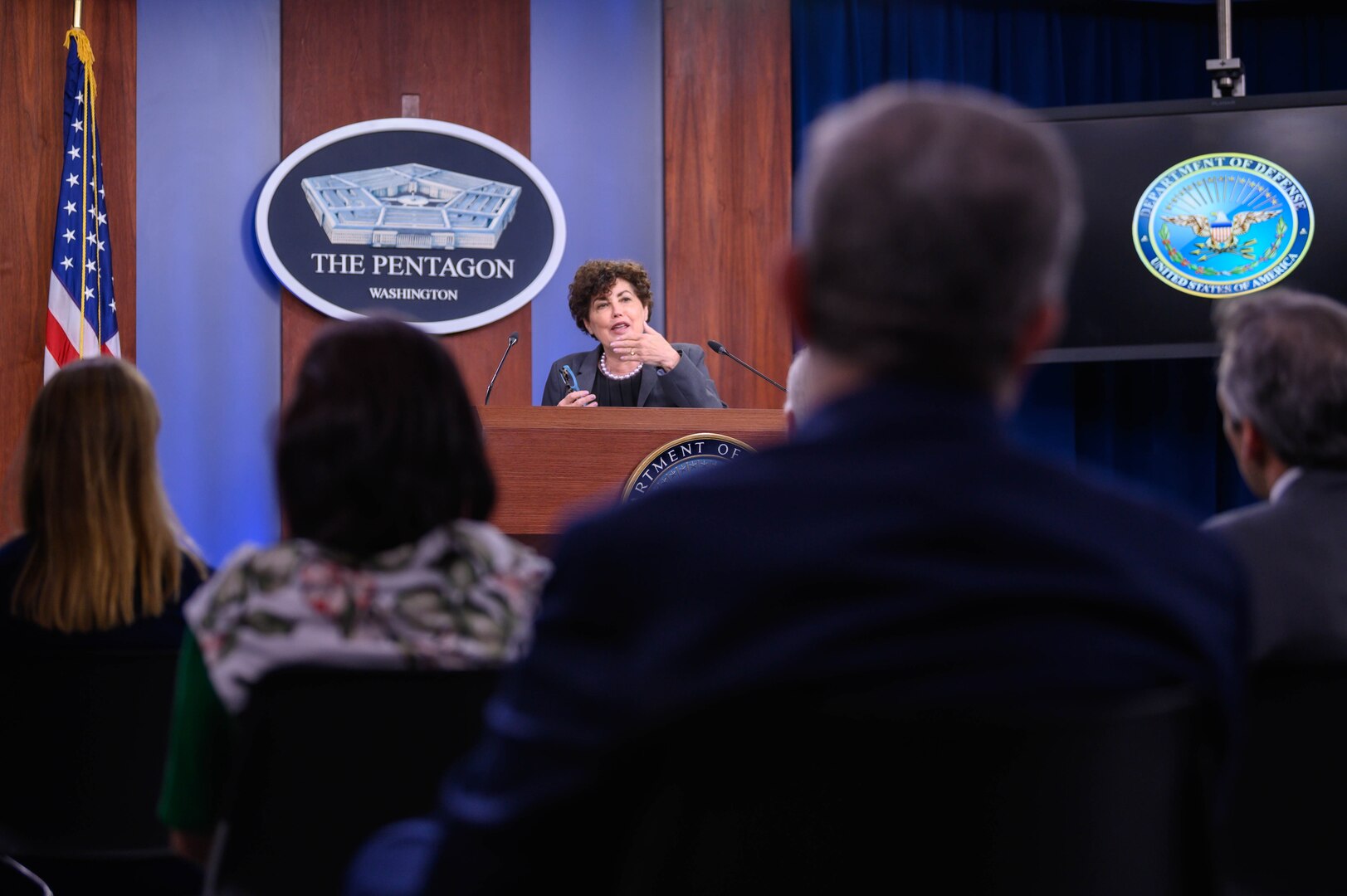 A woman stands behind a lectern. Before her is an audience of reporters.