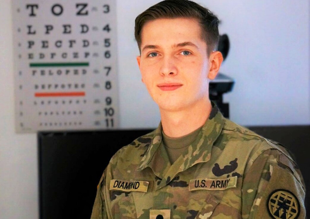 Spc. Andrew Diamond stands in front of a eye chart in a medical clinic.