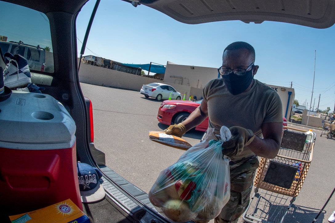 A soldier puts a bag of food into the back of a car.