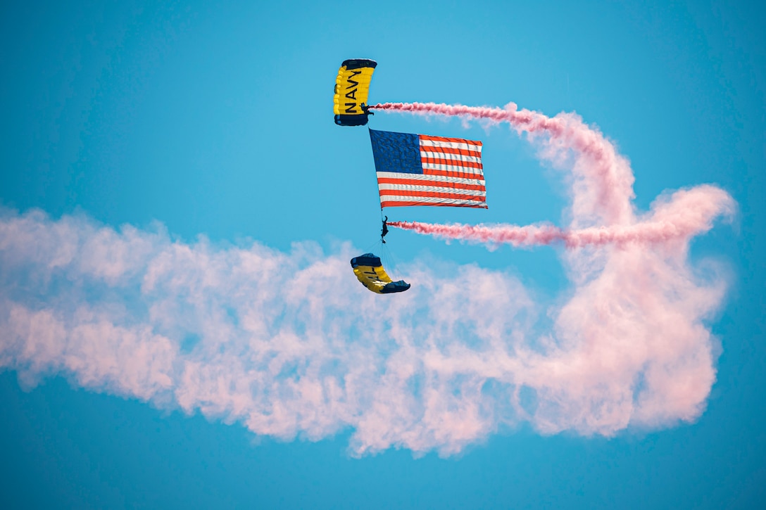 Two sailors display an American flag while freefalling with parachutes and spraying clouds of red smoke into the air.
