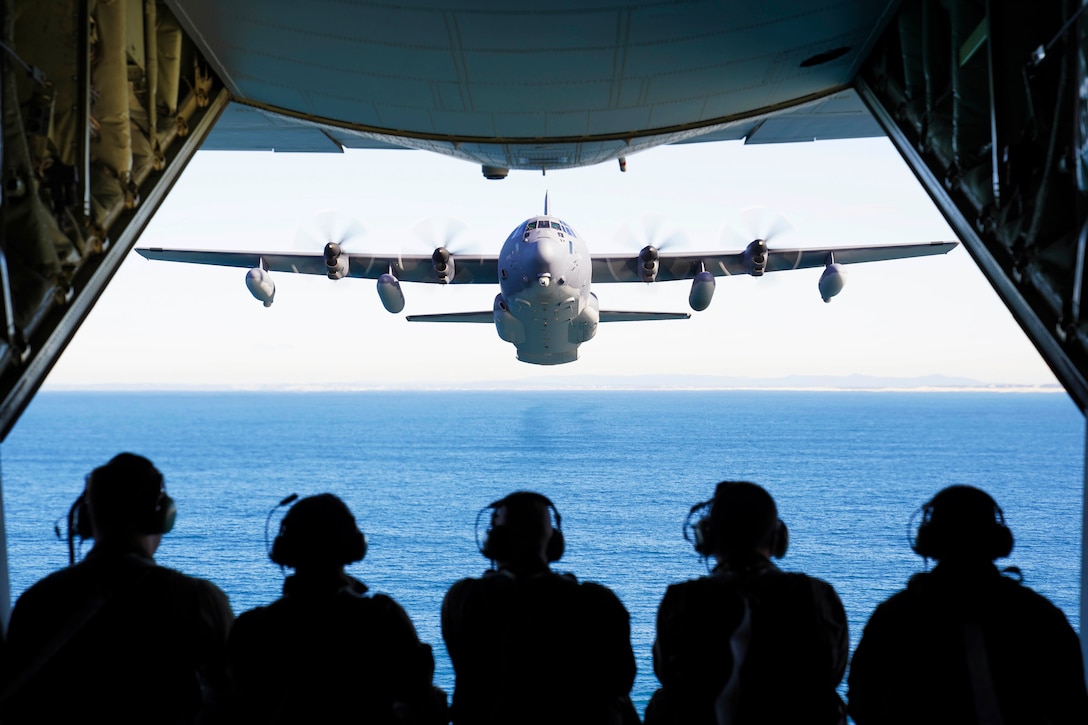 Five airmen sit at the back of an aircraft watching another aircraft fly behind.