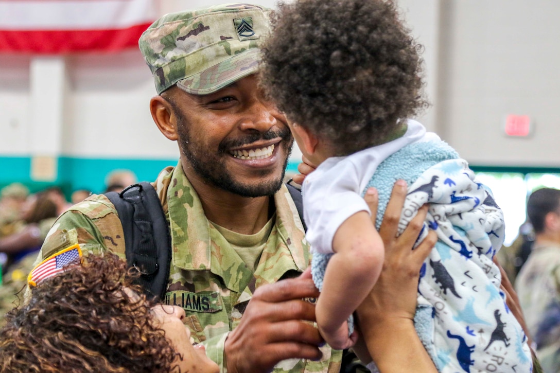 A soldier smiles at a baby that is being held by a woman.