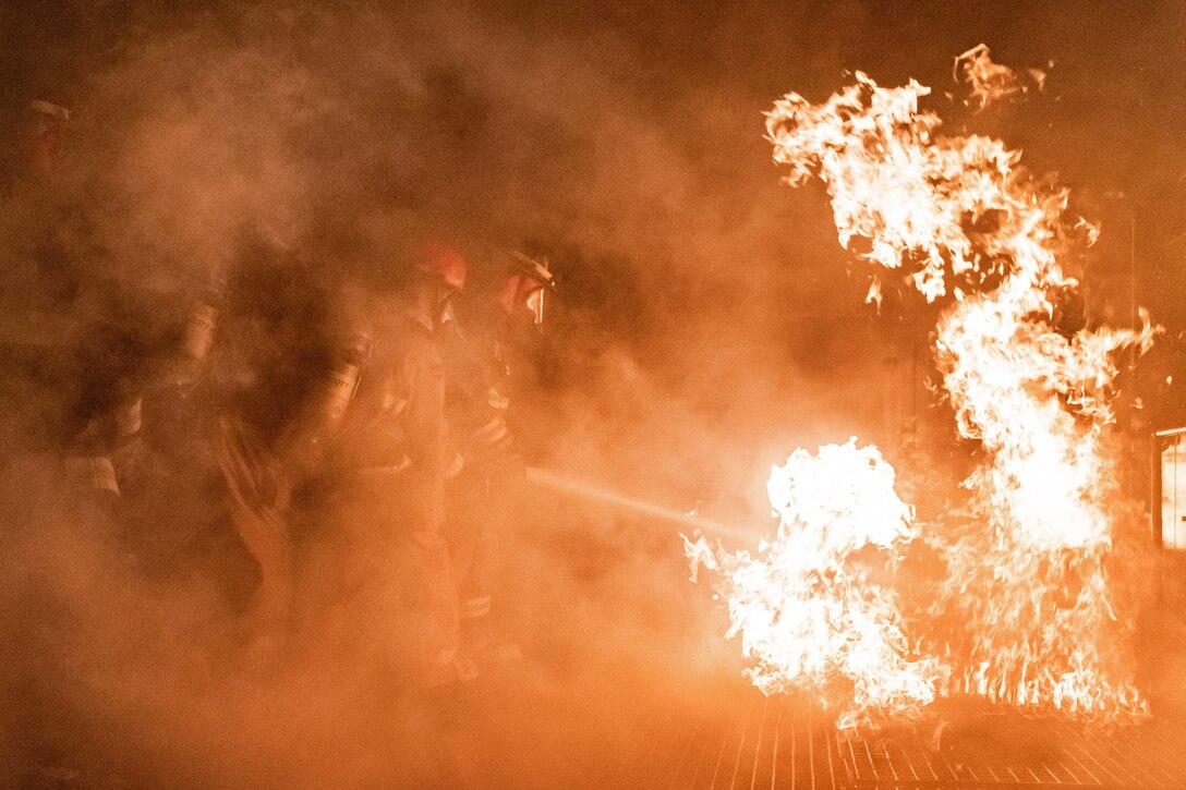 Navy firefighters spray water on flames in an enclosed space as smoke partially obscures them.