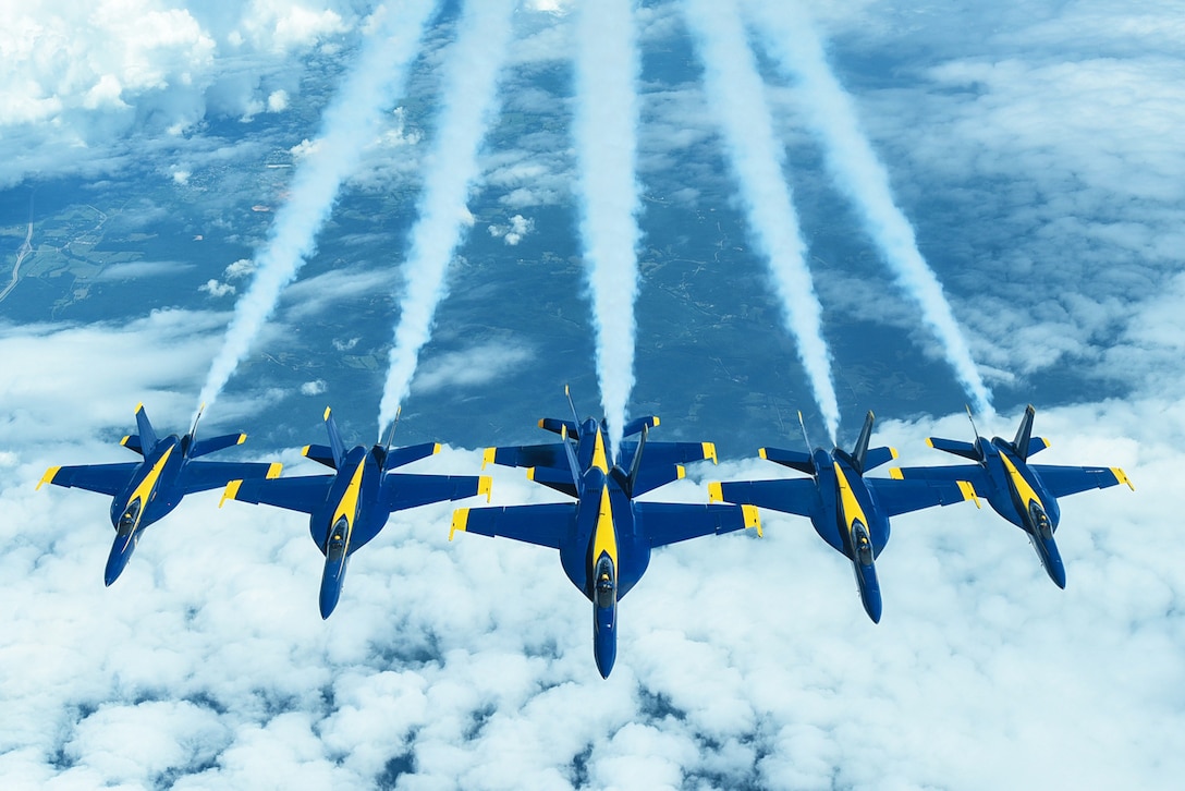 Six Navy aircraft fly in formation in blue sky above clouds, leaving trails in their wake.