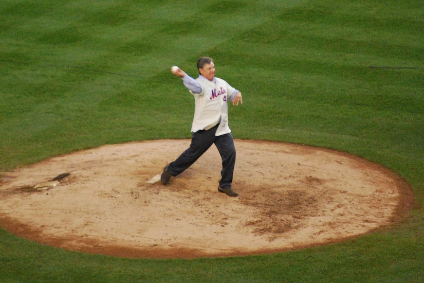 A man dressed in street clothes throws a baseball from the pitcher's mound.