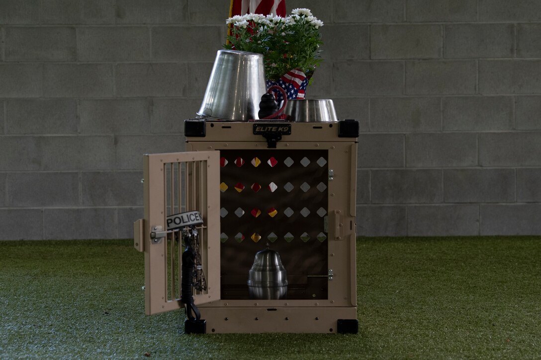 A photo of a dog crate with a food and water bowl, flowers, and a dog toy on top, and an urn inside the crate, and a dog collar and leash on the crate door.