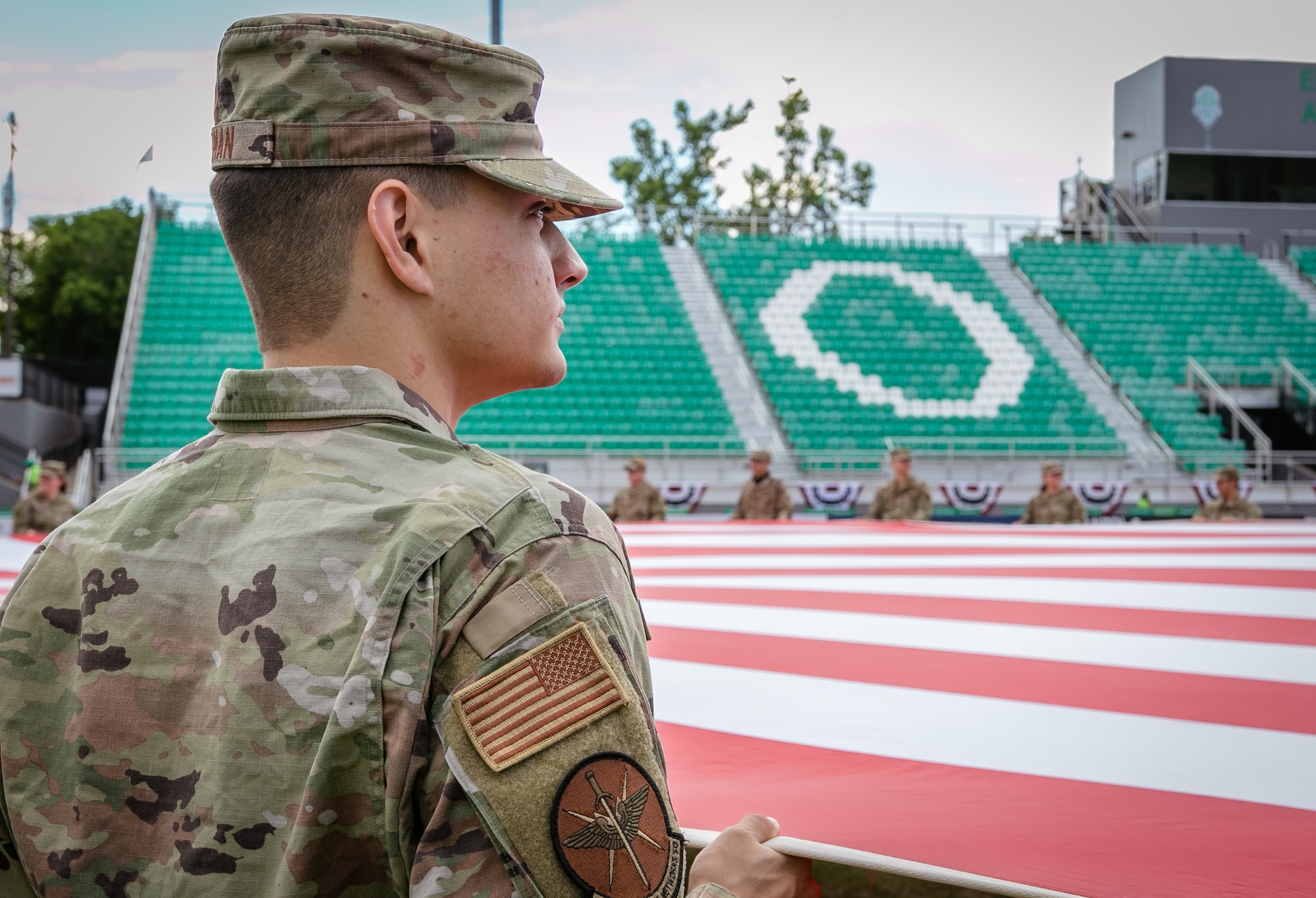 Airman holds section of large U.S. flag at stadium.