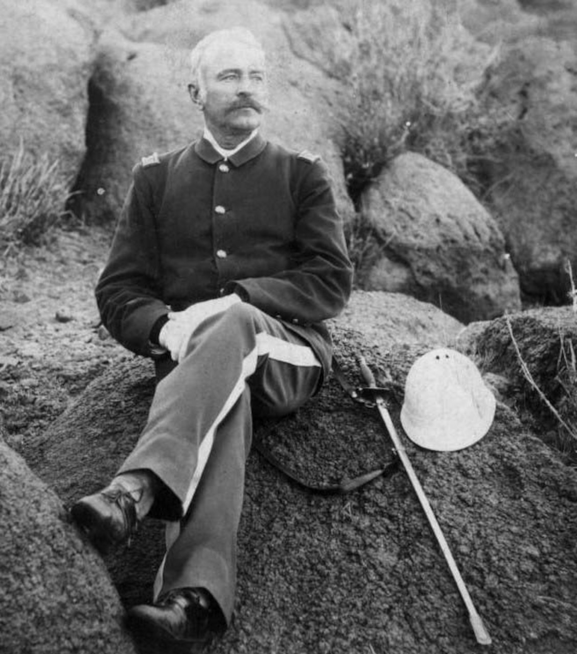 A man sits on a rock with one knee crossed over the other. A scabbard and helmet sit beside him.