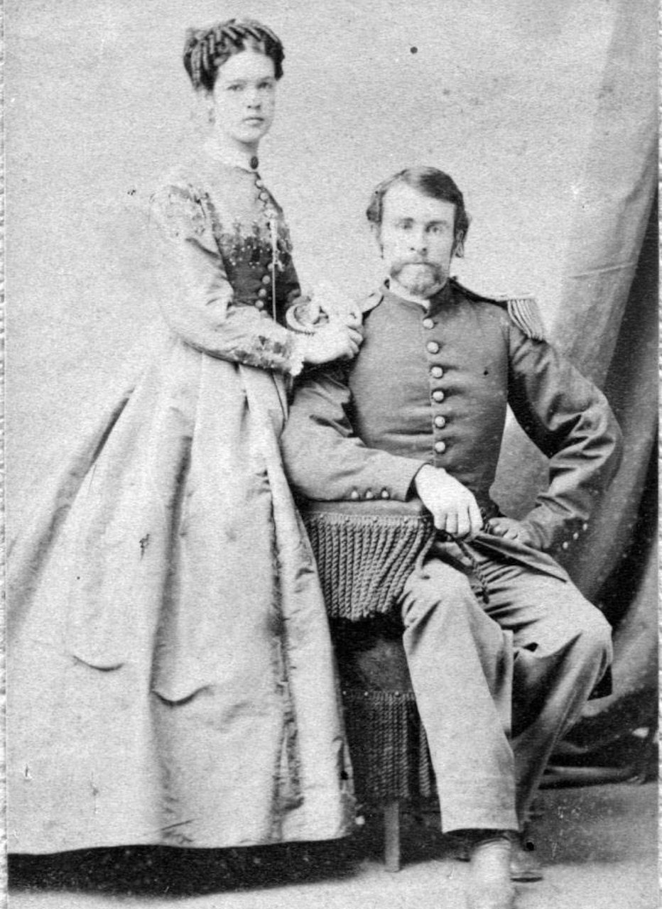 A man in dress uniform sits in a chair. A woman stands beside him, her hand on his shoulder.