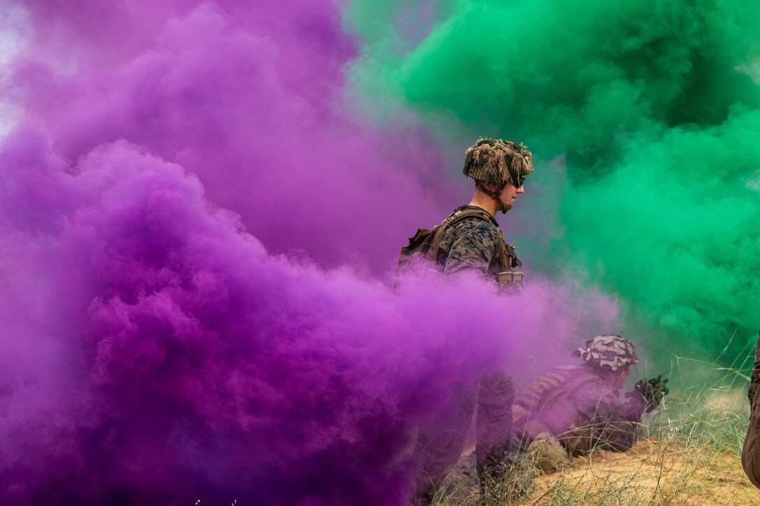 Marines train in a field through purple and green clouds of smoke.