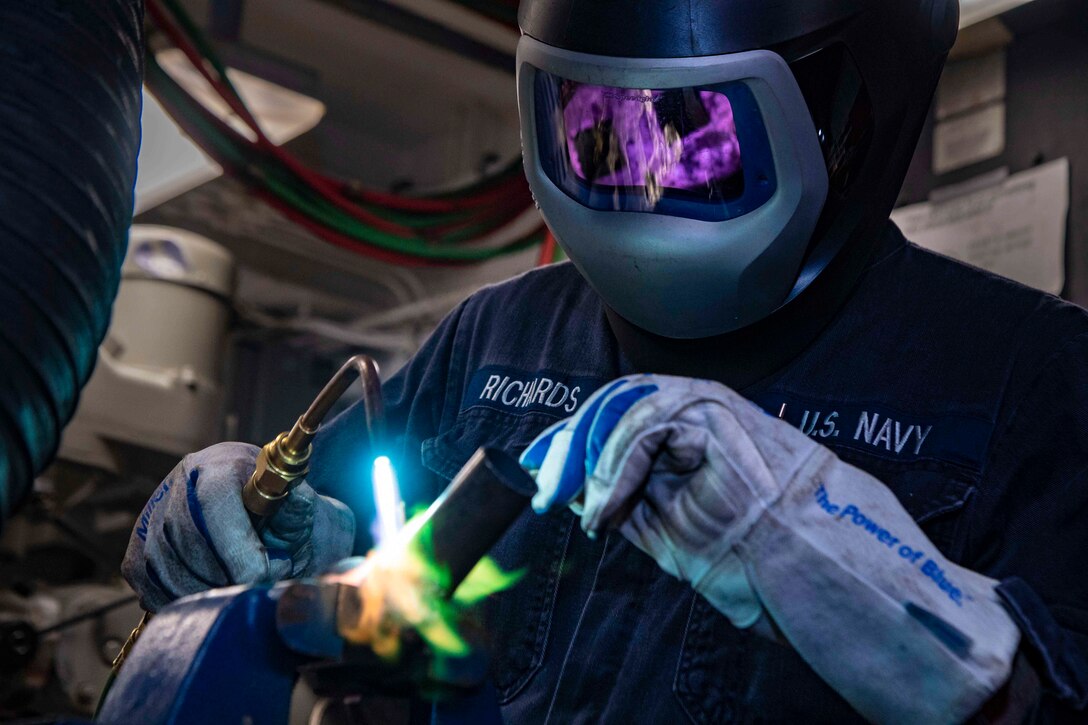 A sailor wearing welding gear uses a small torch on a pipe.