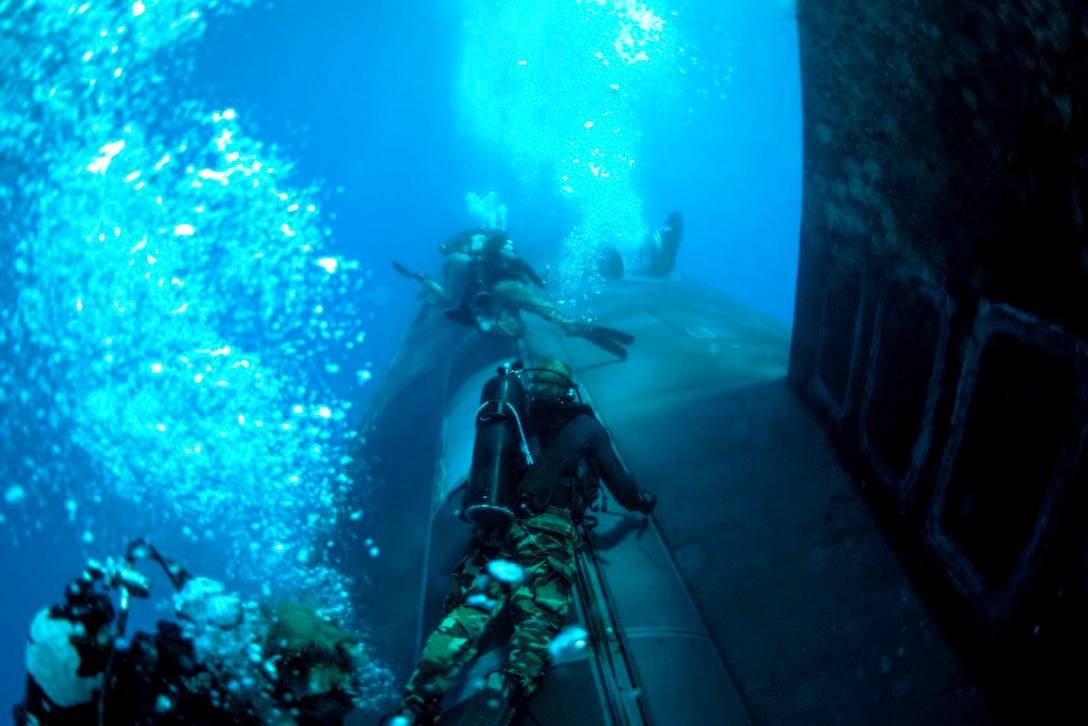 Navy SEALs wearing dive gear swim near a submarine underwater.