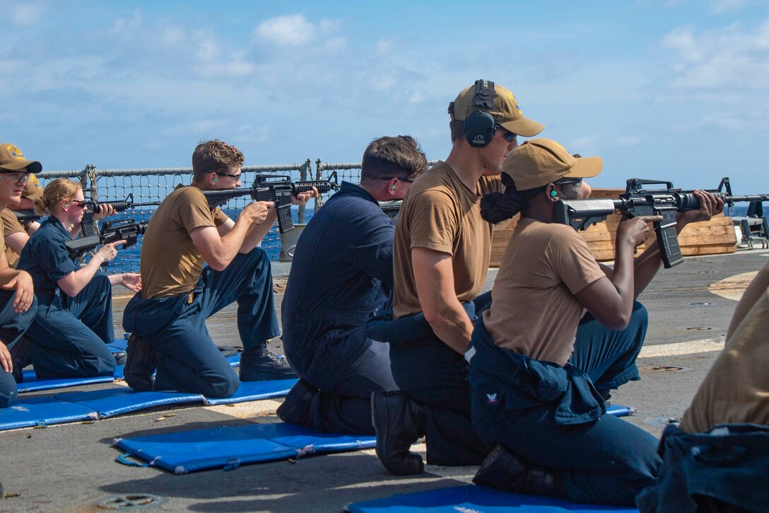 Sailors kneel on the deck of a ship pointing weapons at targets.