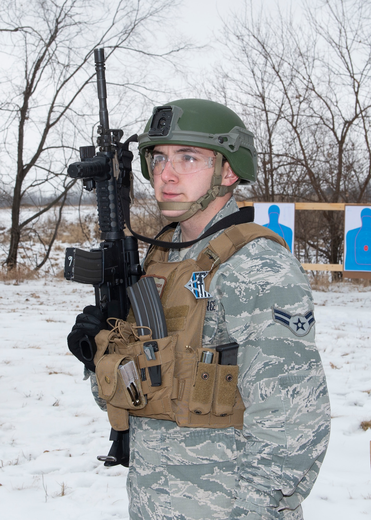 Airman in uniform, wearing protective helmet and standard size tactical vest while holding a weapon in his right hand