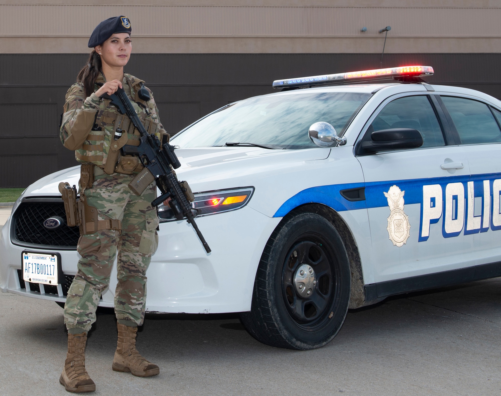 Female Airman in uniform standing guard while holding her weapon, in front of a security forces police car with it's lights flashing. Airman wearing new body armor specifically designed for women