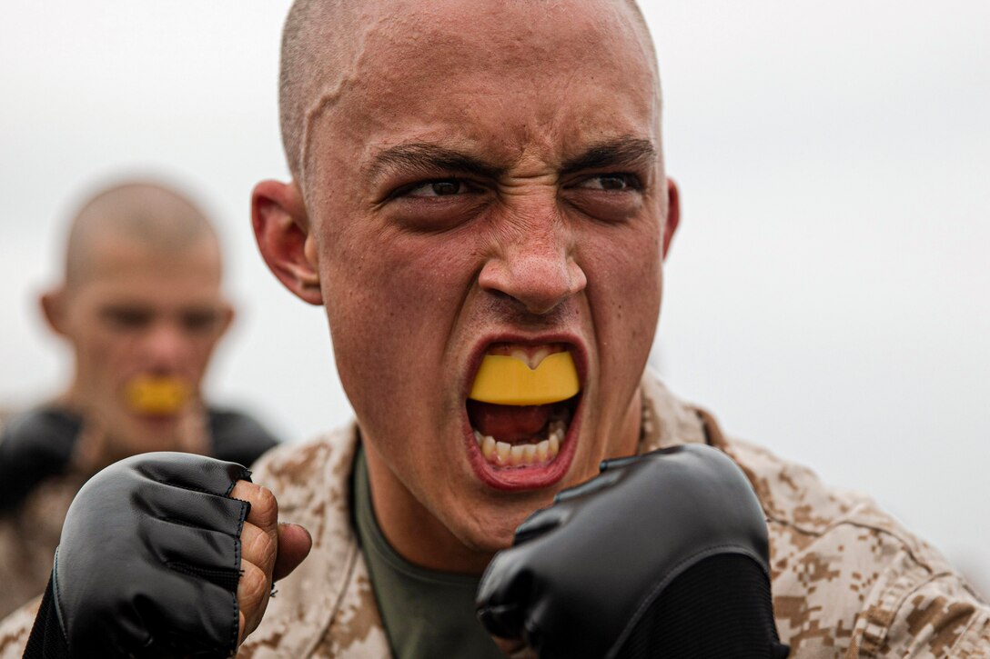 A Marine Corps recruit wearing a mouth guard holds up black-gloved fists and scowls.