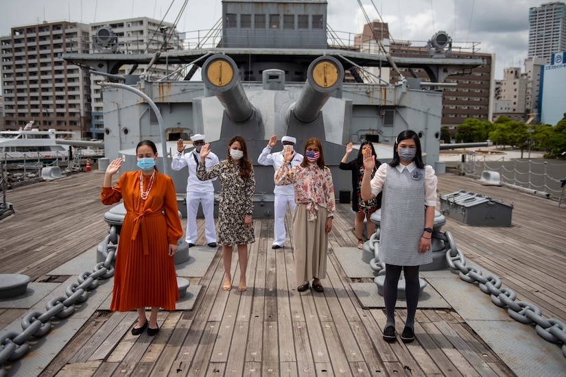 Seven men and women hold up their right hands outdoors on a ship.