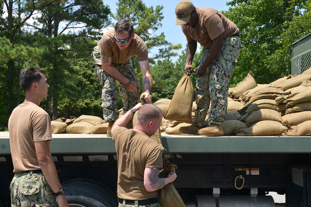 Two sailors stand an open truck bed filled with sandbags, which they pass to two sailors on the ground.