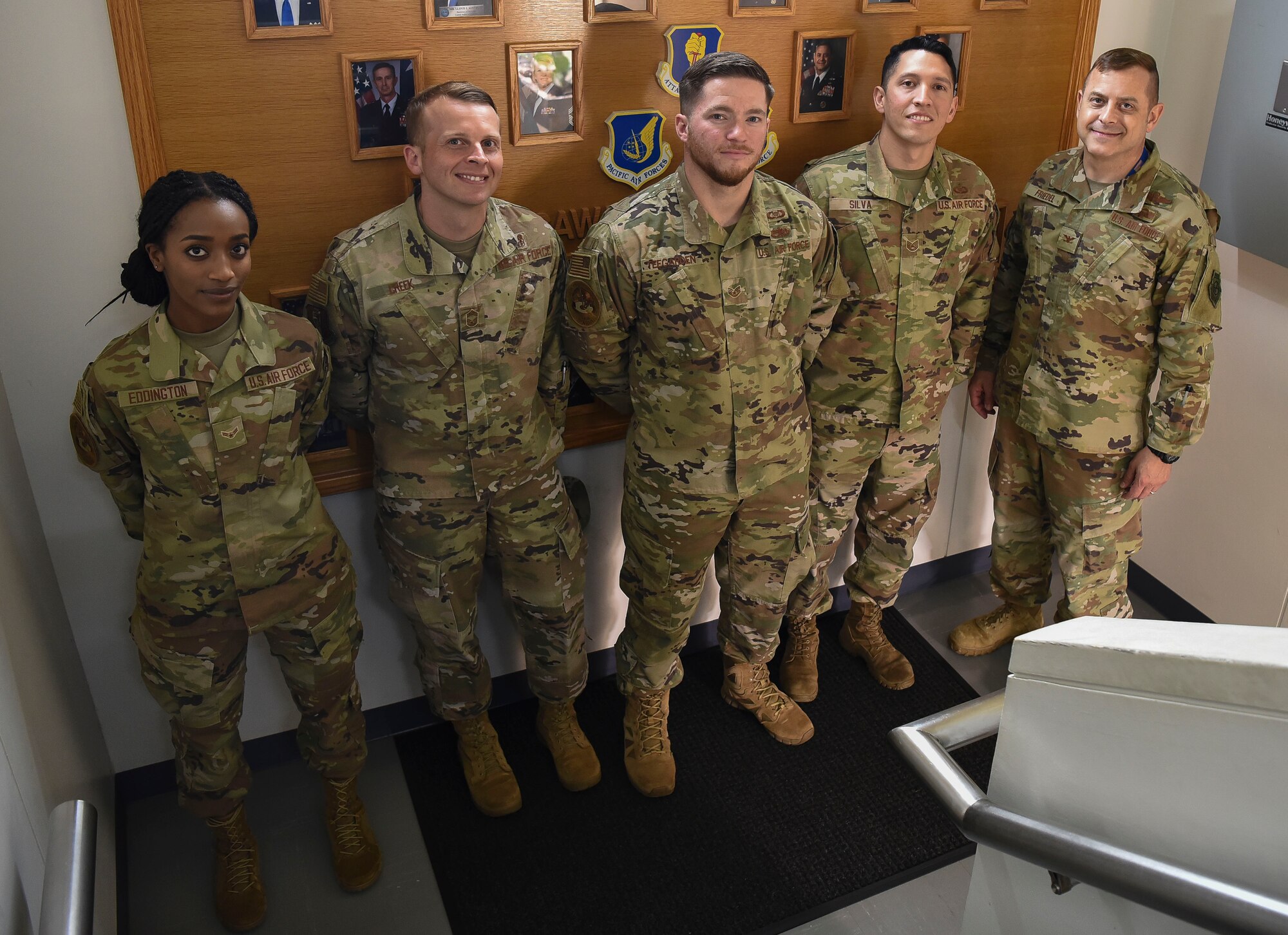 Military members stand side3 by side posing for a picture in front of a leadership photo wall in a stair case.