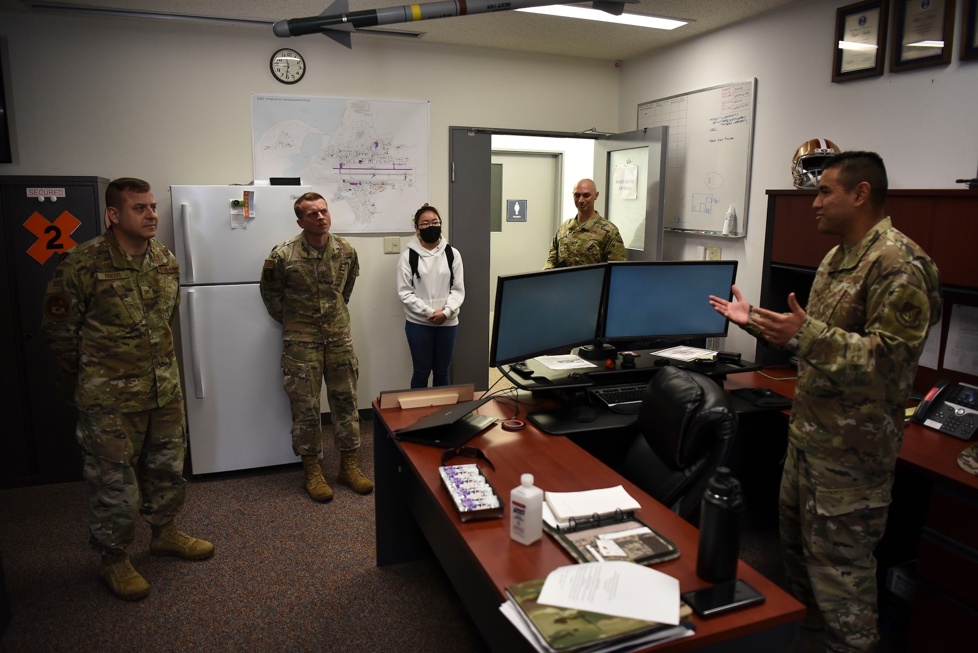 A group of military members surround one speaking behind a desk.