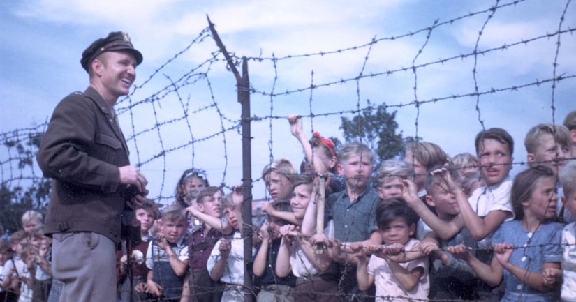 A smiling U.S. military pilot talks to children standing on the other side of a wire fence.