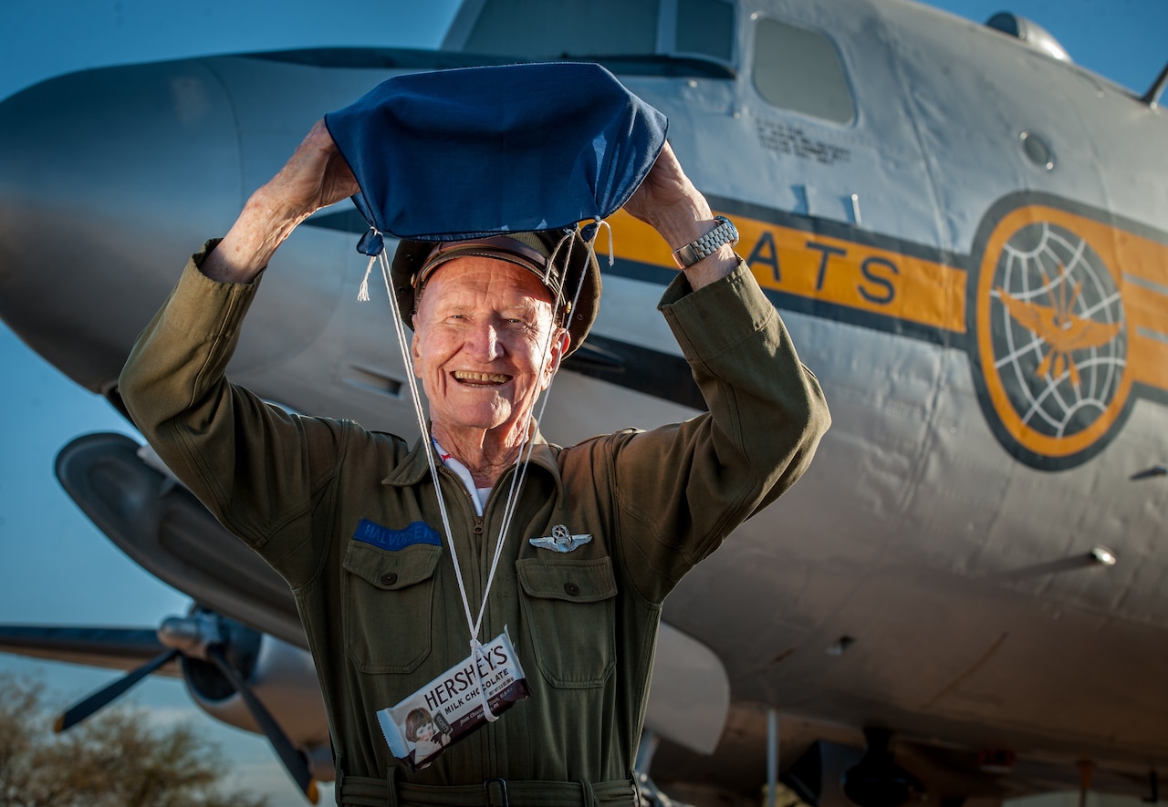 A smiling veteran holds up a small parachute attached to a candy bar while standing in front of an aircraft.