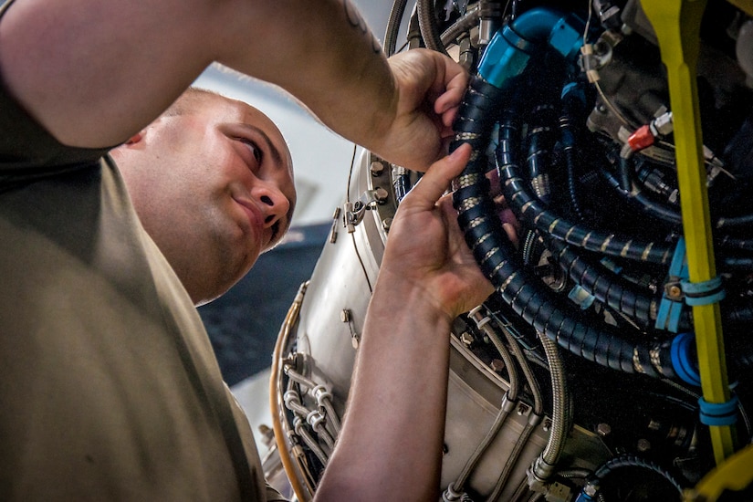 A man in a brown T-shirt adjusts hoses in a military helicopter engine.