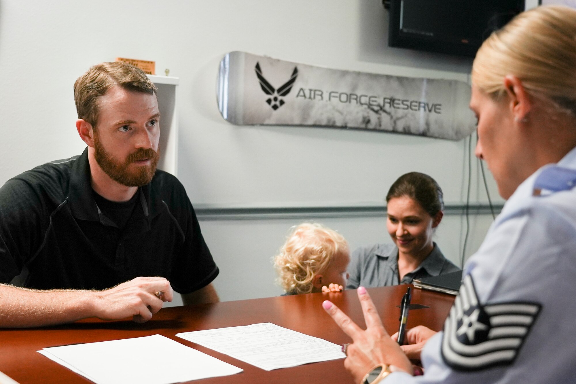 Civilian leans on desk over two documents and asks Airman in blues questions while his partner and child wait in background.